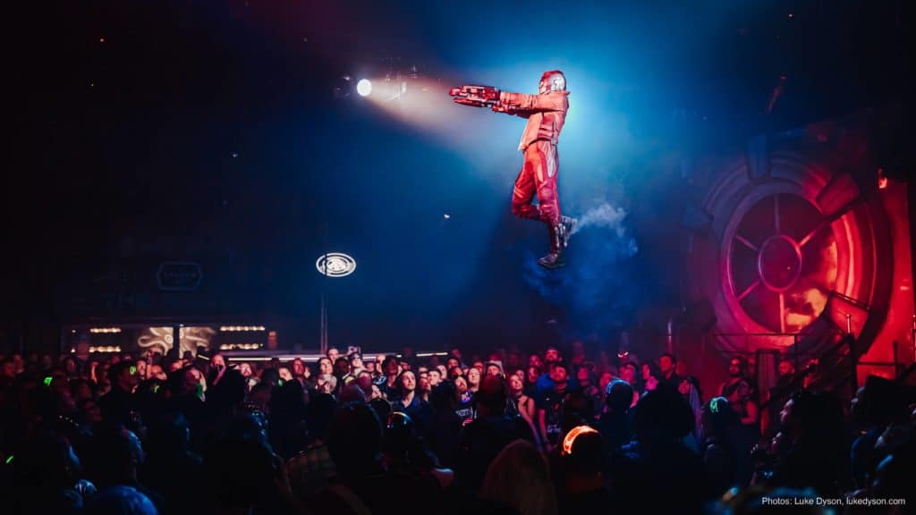 crowds looking up to a suspended person in red costume with smoke coming out of their shoes against a theatre spotlight