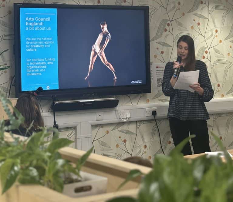 Abigail McQuater standing with long brown hair reading from notes infront of a presentation slide tv with green plants in the foreground